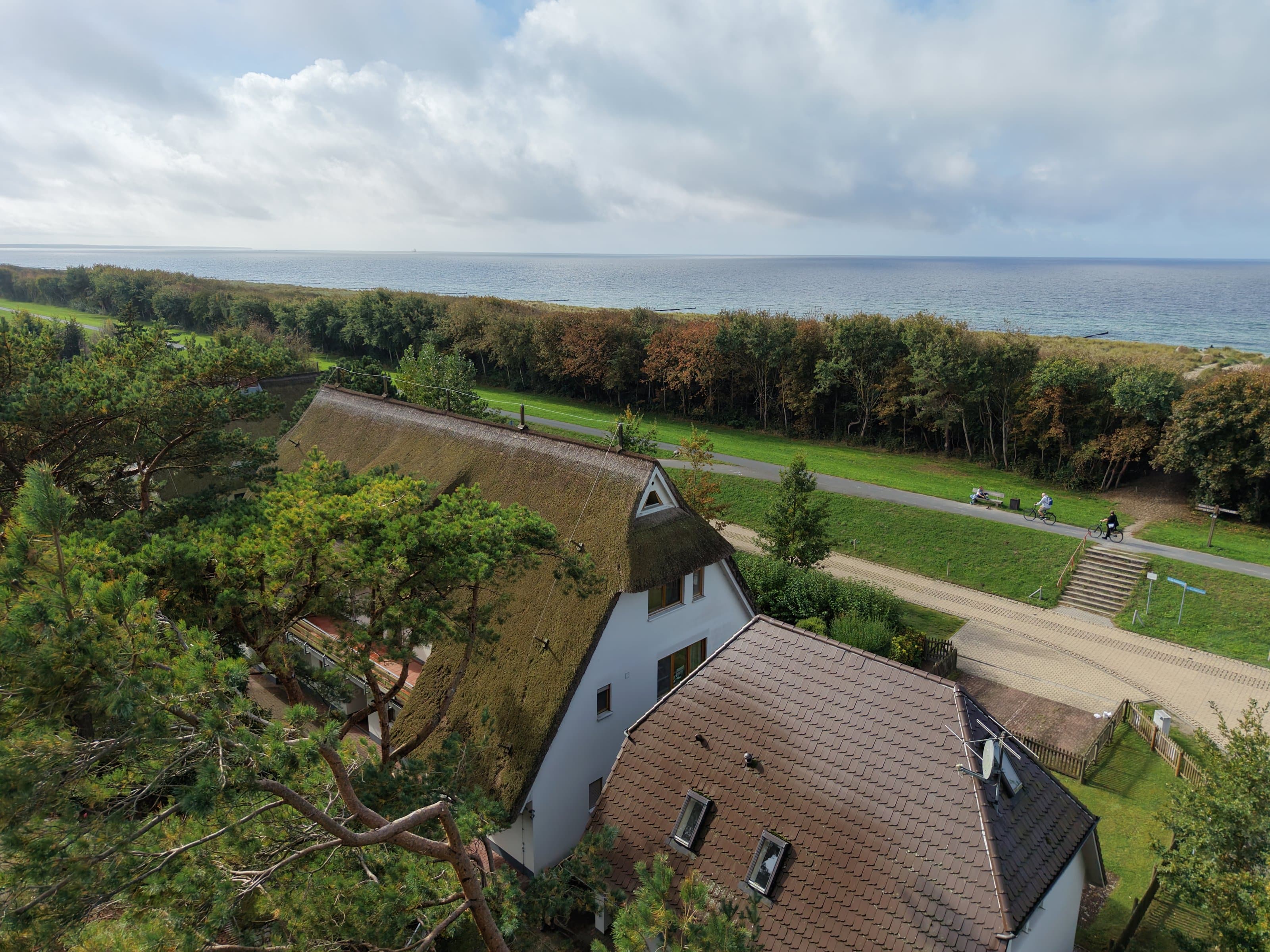 Luftaufnahme am Mittag: weißes Haus mit Reetdach zwischen Kiefern, davor ein Haus mit Ziegeldach, dahinter Straße, Deich mit Treppe und Radweg, Gehölzstreifen und die Ostsee unter Wolken