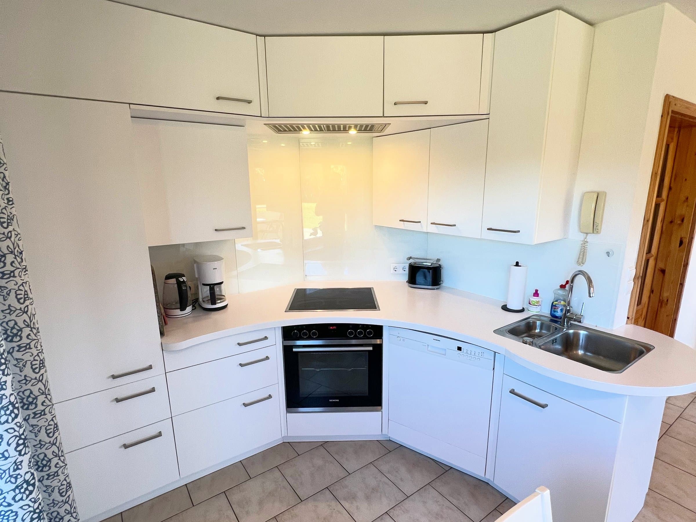 Weiße Eckküche mit Kochfeld, Backofen, Geschirrspüler, Spüle, Kaffeemaschine, Wasserkocher und Toaster